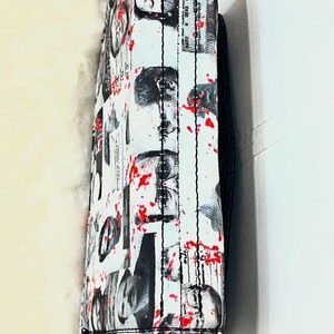 May include: A white rectangular bag featuring a black and white collage of human faces and newspaper-style text, accented with scattered red paint splatters. The bag has black stitched seams and a black strap attached to the top edge.