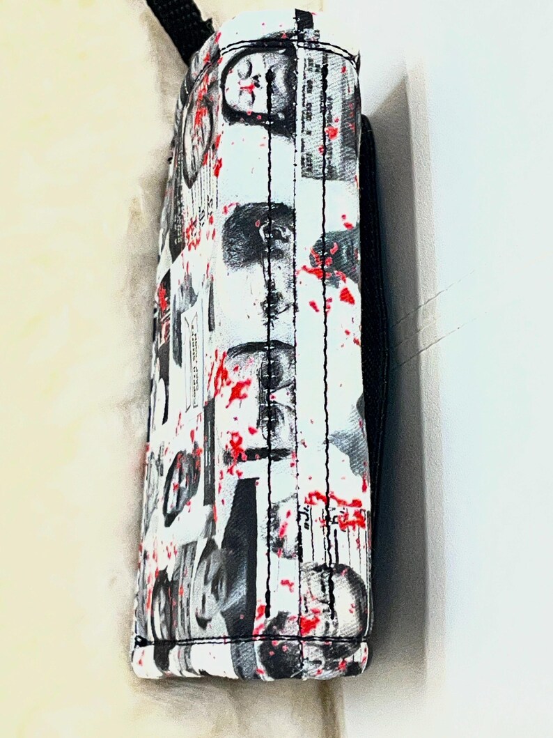 May include: A white rectangular bag featuring a black and white collage of human faces and newspaper-style text, accented with scattered red paint splatters. The bag has black stitched seams and a black strap attached to the top edge.