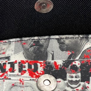 May include: A black wallet with a silver magnetic snap closure. The interior lining features a black and white graphic print of various faces and the word WANTED, accented with scattered red paint splatters across the fabric surface.