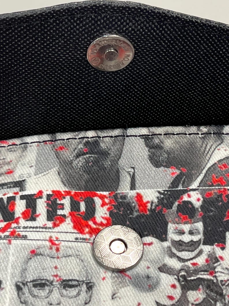 May include: A black wallet with a silver magnetic snap closure. The interior lining features a black and white graphic print of various faces and the word WANTED, accented with scattered red paint splatters across the fabric surface.