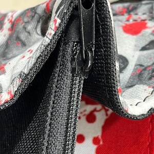 May include: A close-up view of a black zipper mechanism on a white fabric bag featuring a red and black splatter pattern. The zipper pull is black and metallic, integrated into a black fabric seam that connects the patterned panels of the bag.