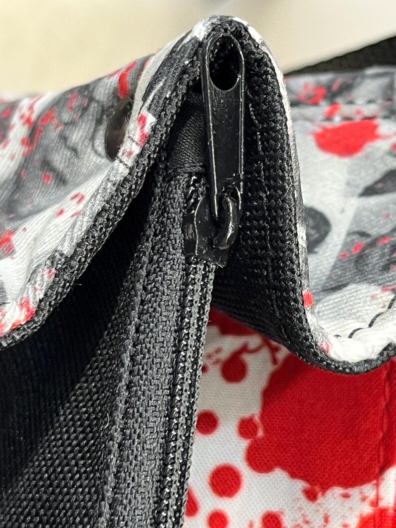 May include: A close-up view of a black zipper mechanism on a white fabric bag featuring a red and black splatter pattern. The zipper pull is black and metallic, integrated into a black fabric seam that connects the patterned panels of the bag.