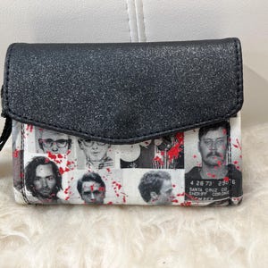 May include: A black glittery wristlet wallet featuring a collage of black and white mugshot portraits overlaid with red paint splatters. The wallet has a fold-over flap closure and a black wrist strap. One portrait includes text reading 4 28 73 29076 SANTA CRUZ CO SHERIFF CORONER.
