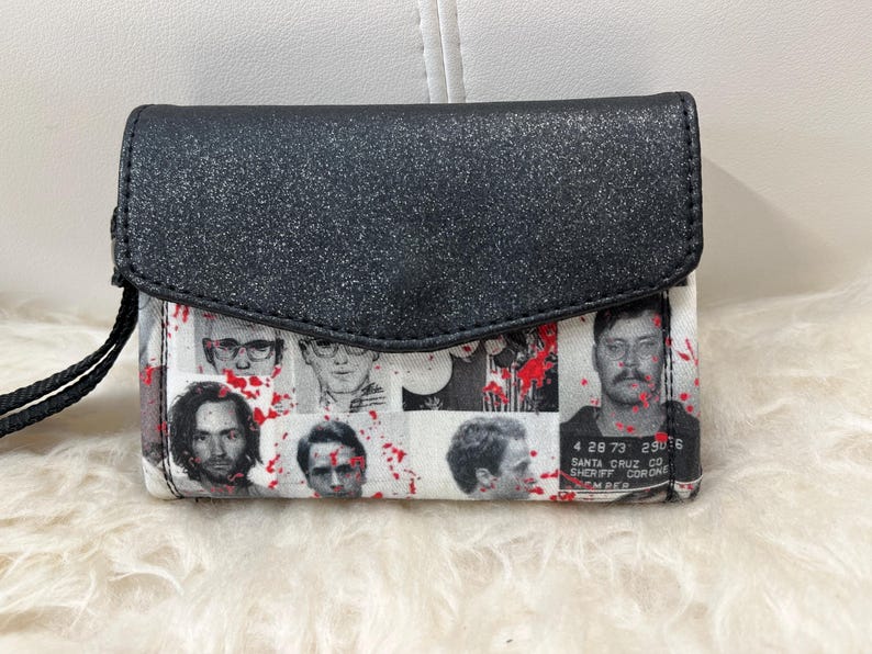 May include: A black glittery wristlet wallet featuring a collage of black and white mugshot portraits overlaid with red paint splatters. The wallet has a fold-over flap closure and a black wrist strap. One portrait includes text reading 4 28 73 29076 SANTA CRUZ CO SHERIFF CORONER.
