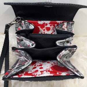 May include: A black wristlet wallet with an open interior featuring a white lining printed with red blood splatter patterns and grayscale portraits of people. The wallet includes multiple card slots, a central zippered pocket, and a black wrist strap.