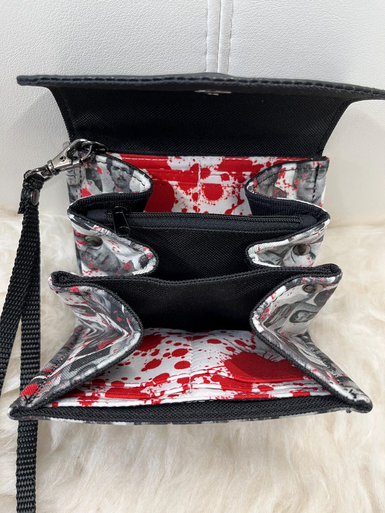 May include: A black wristlet wallet with an open interior featuring a white lining printed with red blood splatter patterns and grayscale portraits of people. The wallet includes multiple card slots, a central zippered pocket, and a black wrist strap.