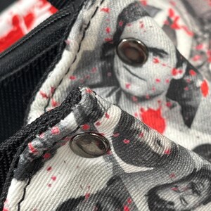 May include: A fabric garment featuring a black and white photographic print of faces overlaid with scattered red paint splatters. The item includes black trim along the edges and two metallic, circular snap buttons secured on the textured material.