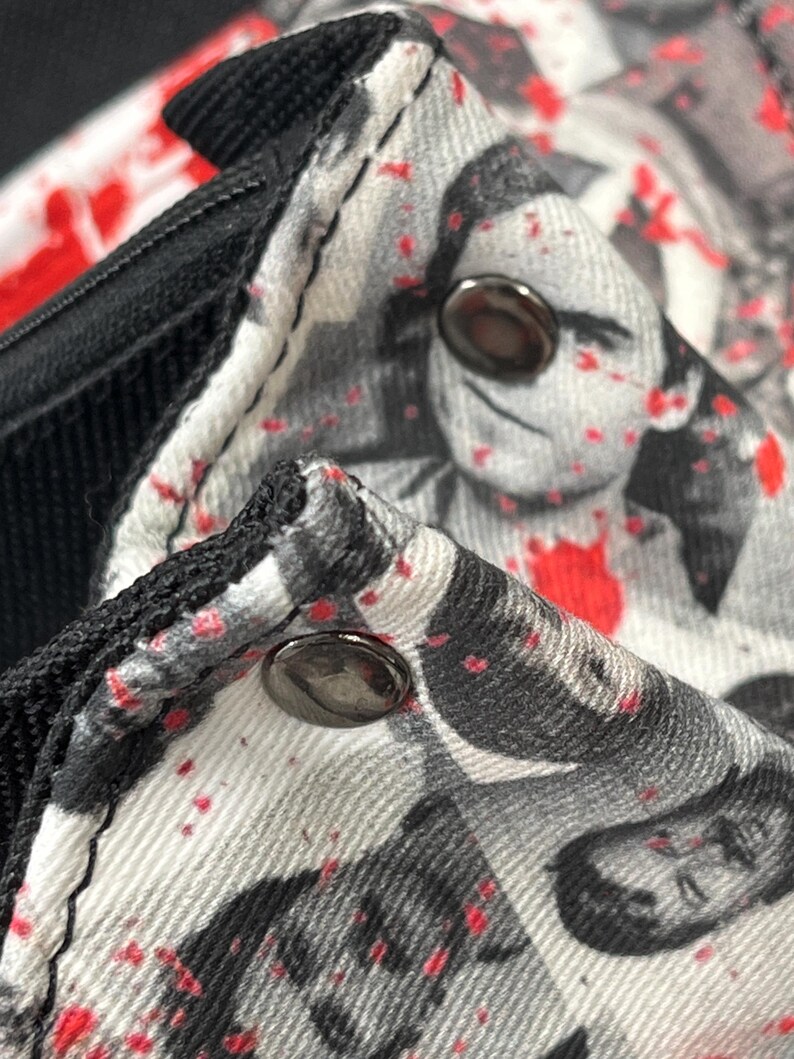 May include: A fabric garment featuring a black and white photographic print of faces overlaid with scattered red paint splatters. The item includes black trim along the edges and two metallic, circular snap buttons secured on the textured material.
