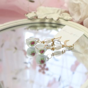 May include: Dangling earrings featuring miniature porcelain teacups with pink rose designs, accented with gold trim. The earrings are connected by faux pearls and gold-colored metal. A tag with the text "Lauren Rose" is visible.