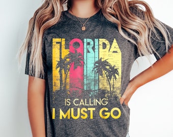 May include: A grey t-shirt with a vintage-style graphic that says "Florida is calling I must go". The graphic features palm trees and a colourful striped background.