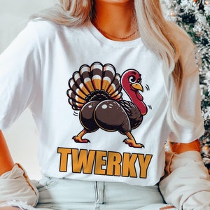 Könnte beinhalten: Weißes T-Shirt mit einem Cartoon-Truthahn-Design. Der Truthahn hat einen braunen Körper, einen roten Kopf und einen großen, bunten Schwanz. Das Wort "TWERKY" ist in Orange darunter gedruckt. Ein humorvolles, lässiges Design.