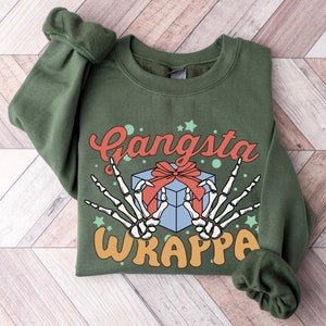 Könnte beinhalten: Olivgrünes Sweatshirt mit den Worten "Gangsta Wrappa" in Orange und Rot. Das Design umfasst eine blaue Geschenkbox mit roter Schleife und zwei Skeletthänden, die ein Friedenszeichen machen.