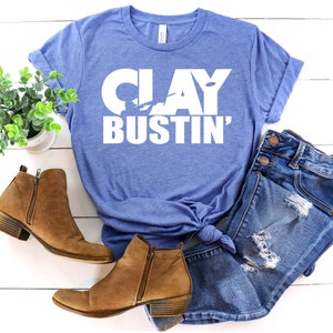May include: A blue t-shirt with the text "CLAY BUSTIN'" in white. The shirt is laid out on a white wooden surface with a pair of brown ankle boots and blue denim jeans.