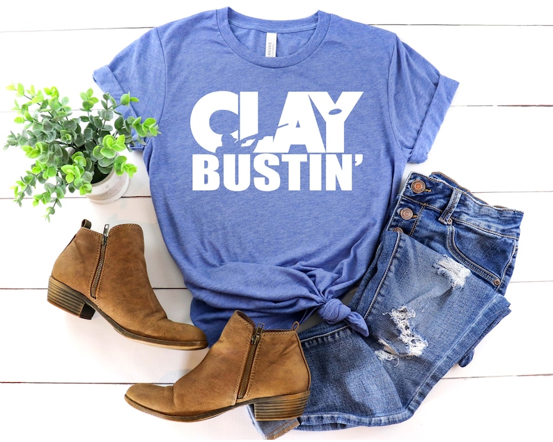May include: A blue t-shirt with the text "CLAY BUSTIN'" in white. The shirt is laid out on a white wooden surface with a pair of brown ankle boots and blue denim jeans.