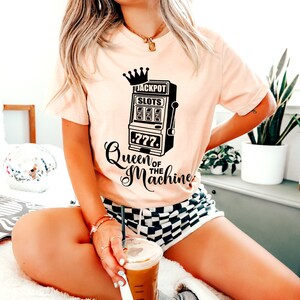 May include: A pink t-shirt with a black graphic of a slot machine with a crown on top and the text "Jackpot Slots 777 Queen of the Machine".