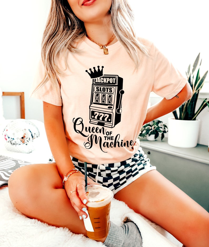 May include: A pink t-shirt with a black graphic of a slot machine with a crown on top and the text "Jackpot Slots 777 Queen of the Machine".