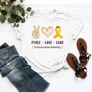 May include: White t-shirt with a peace sign, heart, and yellow ribbon. The text "Peace Love Cure" is below the symbols. The text "Childhood Cancer Awareness" is below the text.