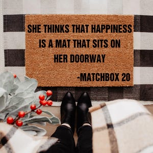 Puede incluir: Una alfombra rectangular marrón con el texto "SHE THINKS THAT HAPPINESS IS A MAT THAT SITS ON HER DOORWAY -MATCHBOX 20" en negro. La alfombra está sobre una alfombra a cuadros en blanco y negro. Se ven botas negras.