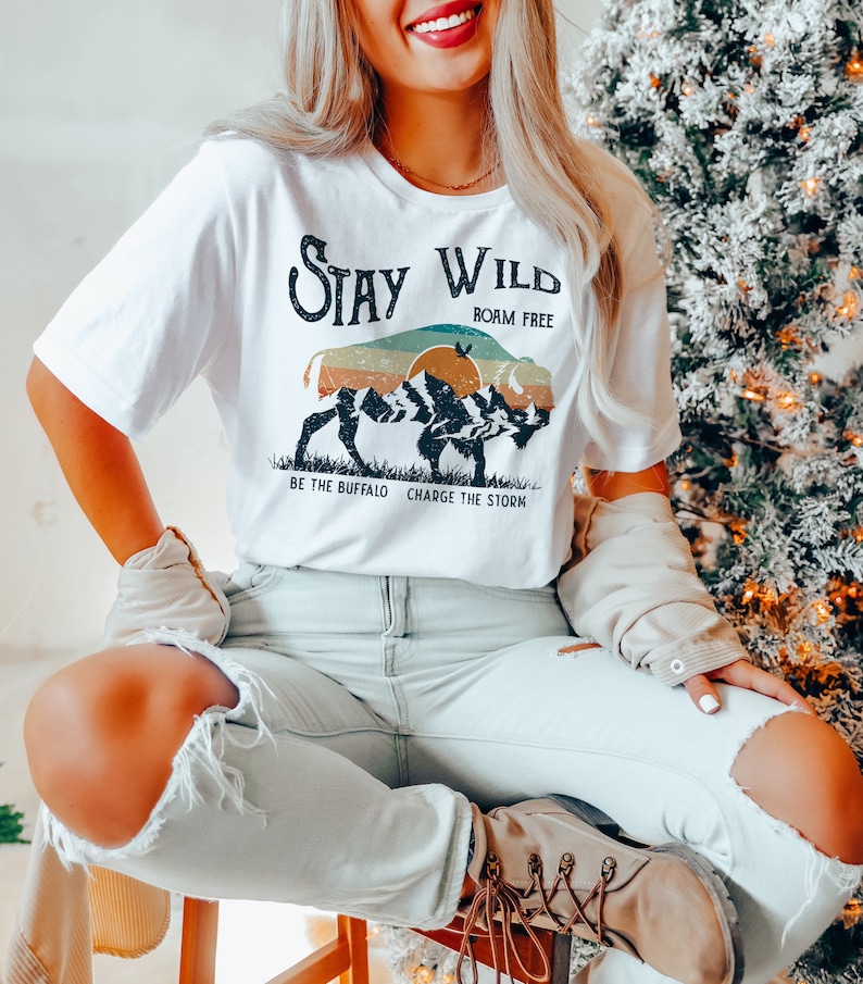 May include: White t-shirt with a graphic of a buffalo silhouette in front of a sunset and mountains. The text "Stay Wild" is above the buffalo and "Roam Free" is below. The text "Be the Buffalo Charge the Storm" is below the buffalo.