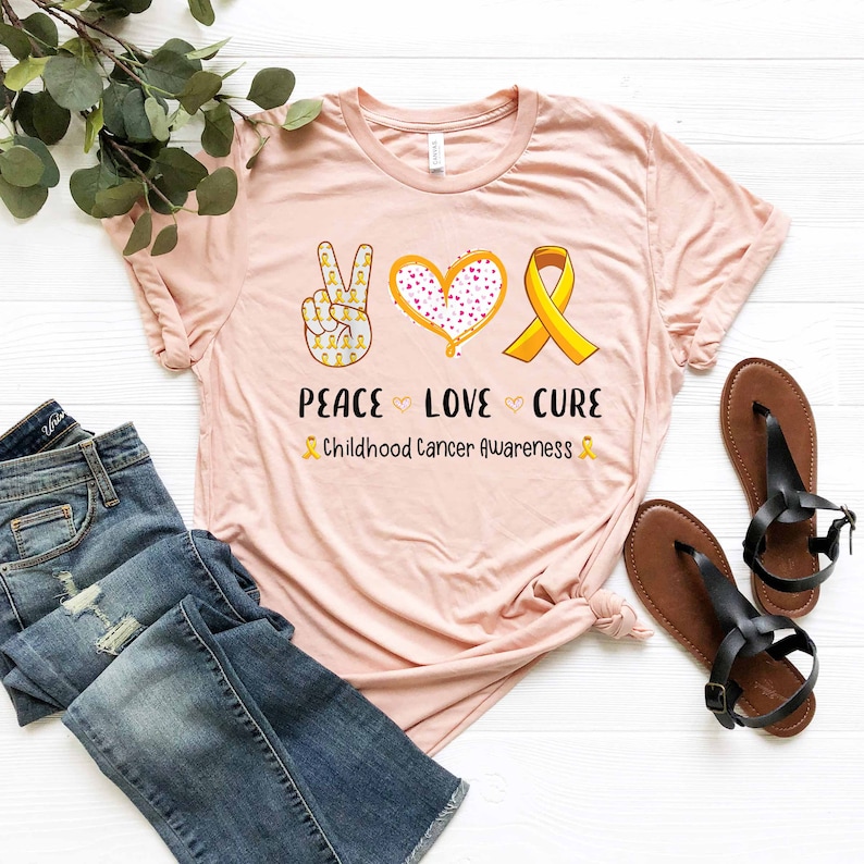 May include: A pink t-shirt with a peace sign, heart, and ribbon design. The peace sign is made of yellow ribbons, the heart is pink with white polka dots, and the ribbon is yellow. The text "Peace Love Cure" is printed below the design, and the text "Childhood Cancer Awareness" is printed below that.