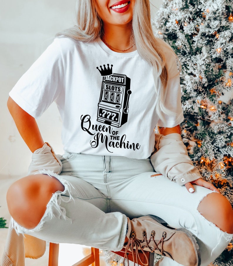 May include: White t-shirt with a black graphic of a slot machine with the text "Jackpot Slots 777 Queen of the Machine".