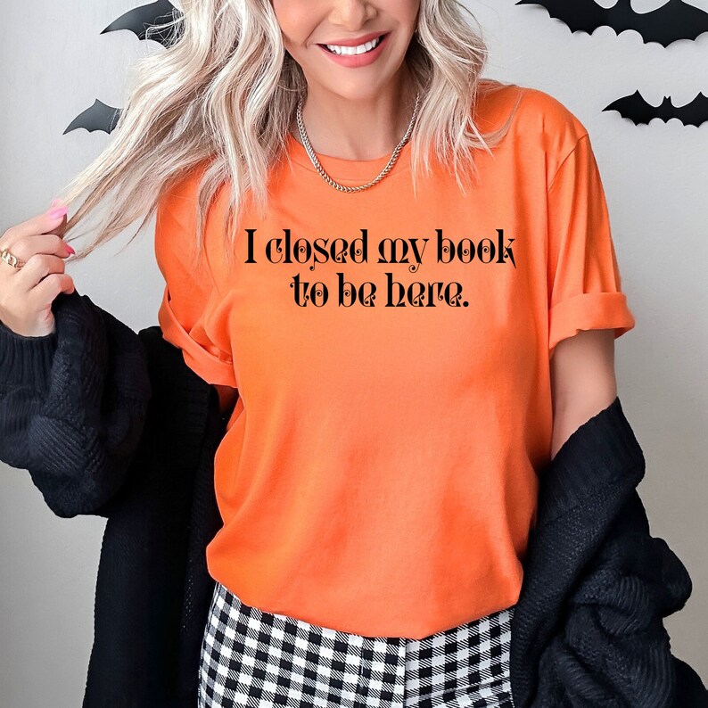 May include: A woman wearing an orange t-shirt with the text "I closed my book to be here." The shirt is a casual style with rolled up sleeves. She is wearing black and white checked trousers and a black cardigan.
