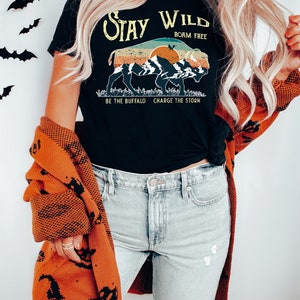 May include: A black t-shirt with a vintage-style graphic of a buffalo silhouette with mountains and a sunset. The text "Stay Wild" is above the buffalo, and "Roam Free" is below. The text "Be the Buffalo Charge the Storm" is at the bottom of the graphic.