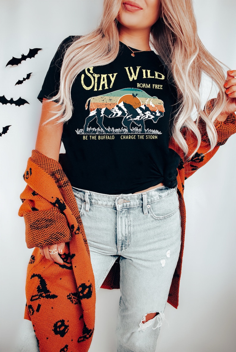 May include: A black t-shirt with a vintage-style graphic of a buffalo silhouette with mountains and a sunset. The text "Stay Wild" is above the buffalo, and "Roam Free" is below. The text "Be the Buffalo Charge the Storm" is at the bottom of the graphic.