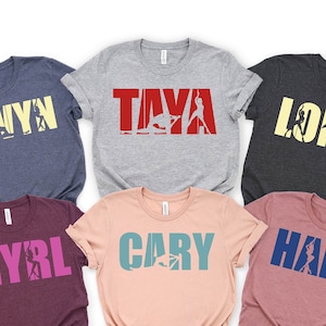May include: Six t-shirts with different names printed on them. The names are "Cwyn", "Taya", "Long", "Myrl", "Cary", and "Hana". Each name is printed in a different color and has a silhouette of a person doing a yoga pose inside the letters.