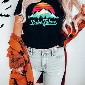 May include: A black t-shirt with a retro sunset graphic and the text "Lake Tahoe" in a blue and white font. The person is wearing a brown cardigan with a black pattern and light blue jeans.