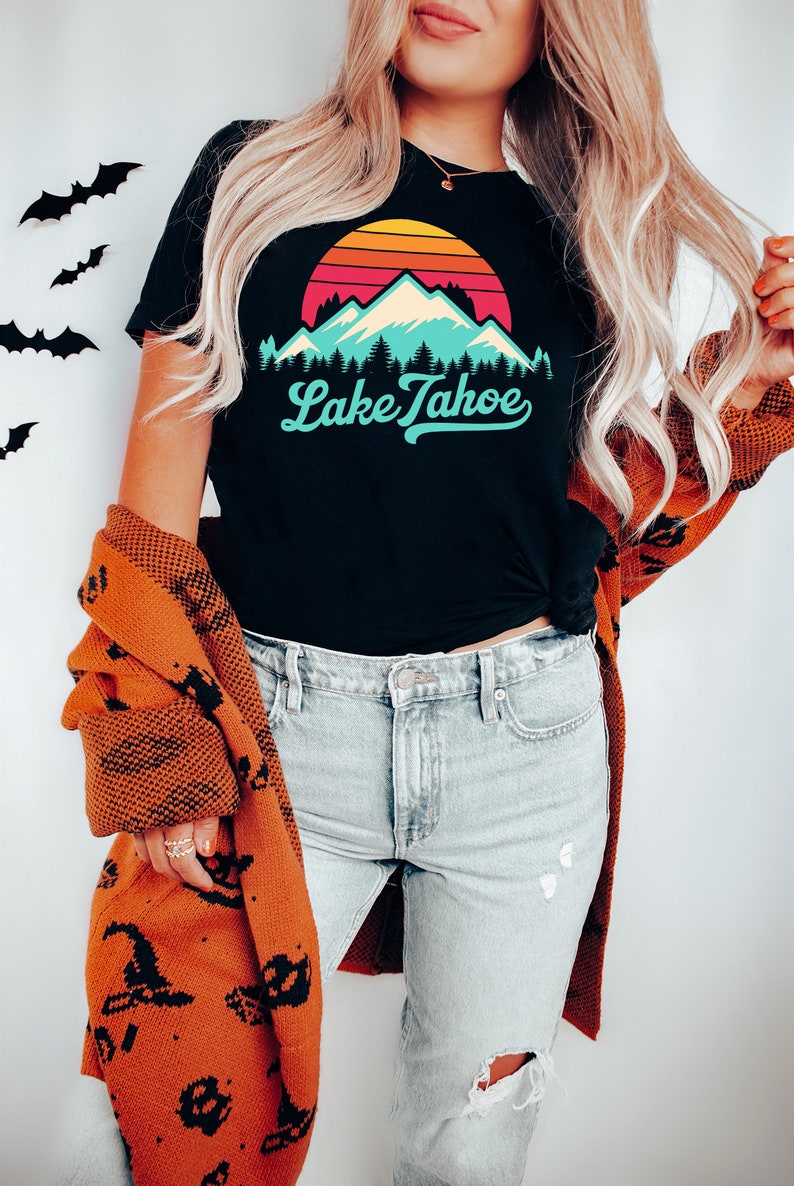 May include: A black t-shirt with a retro sunset graphic and the text "Lake Tahoe" in a blue and white font. The person is wearing a brown cardigan with a black pattern and light blue jeans.