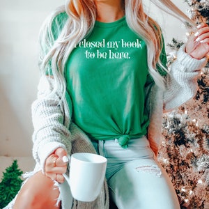 May include: A woman wearing a green t-shirt with the text "I closed my book to be here." She is sitting on a chair with a white mug in her hand. A Christmas tree is in the background.