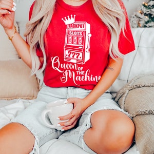 May include: A red t-shirt with a white graphic of a slot machine with the text "Jackpot Slots" and "Queen of the Machine".