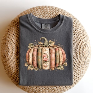 May include: A charcoal gray crew neck t-shirt featuring a graphic print of a pumpkin constructed from vintage-style books with floral patterns and gold accents. The books are arranged in a rounded shape with green leaves at the base and top.