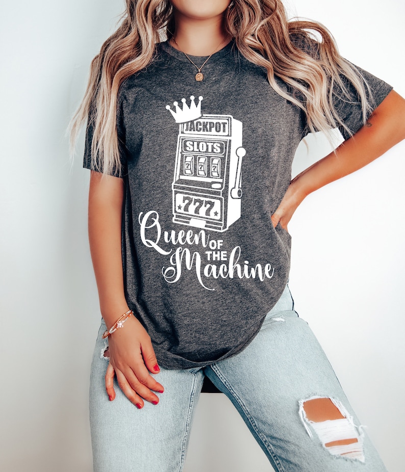 May include: A gray t-shirt with a white graphic of a slot machine with the text "Jackpot Slots" and "Queen of the Machine".