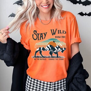 May include: Orange t-shirt with a vintage-style graphic of a buffalo in front of a mountain range with a sunset. The text "Stay Wild Roam Free" is above the buffalo and "Be the Buffalo Charge the Storm" is below the buffalo.