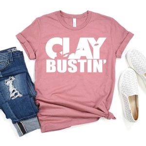 May include: A dusty rose t-shirt with the text "CLAY BUSTIN'" in white lettering. The shirt features a silhouette of a person holding a clay pigeon.