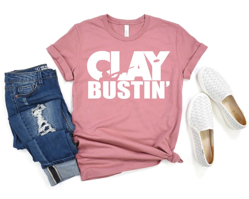 May include: A dusty rose t-shirt with the text "CLAY BUSTIN'" in white lettering. The shirt features a silhouette of a person holding a clay pigeon.
