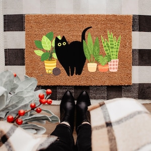 Könnte beinhalten: Eine braune Kokos-Fußmatte mit einer Illustration einer schwarzen Katze und Topfpflanzen. Eine Wollkugel liegt neben der Katze. Die Matte liegt auf einem schwarz-weiß karierten Teppich.