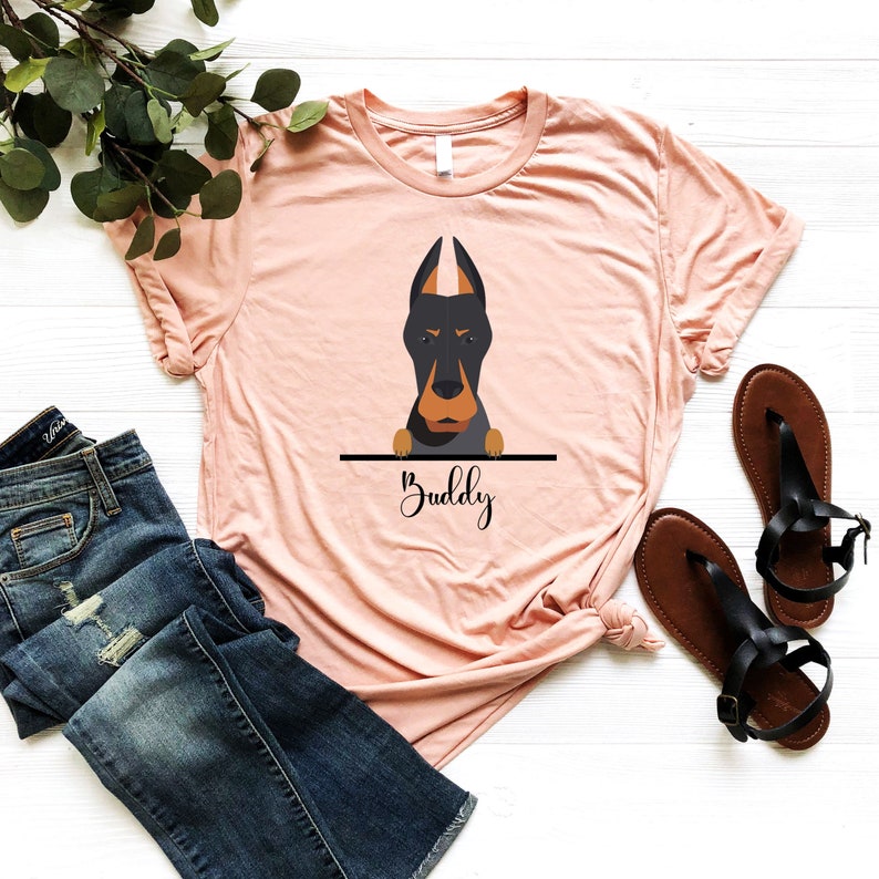 Peut inclure: Un t-shirt couleur p&ecirc;che avec une illustration de chien Dobermann noir et brun qui regarde par-dessus une ligne noire avec le mot "Buddy" &eacute;crit en dessous.