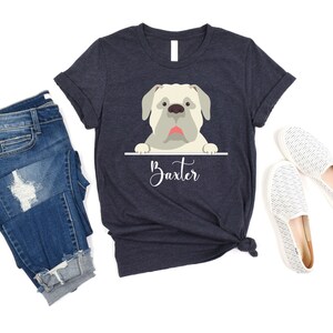 Peut inclure: Un t-shirt bleu marine avec une image de dessin anim&eacute; d'un chien blanc qui regarde par-dessus le haut d'une pancarte qui dit "Baxter".