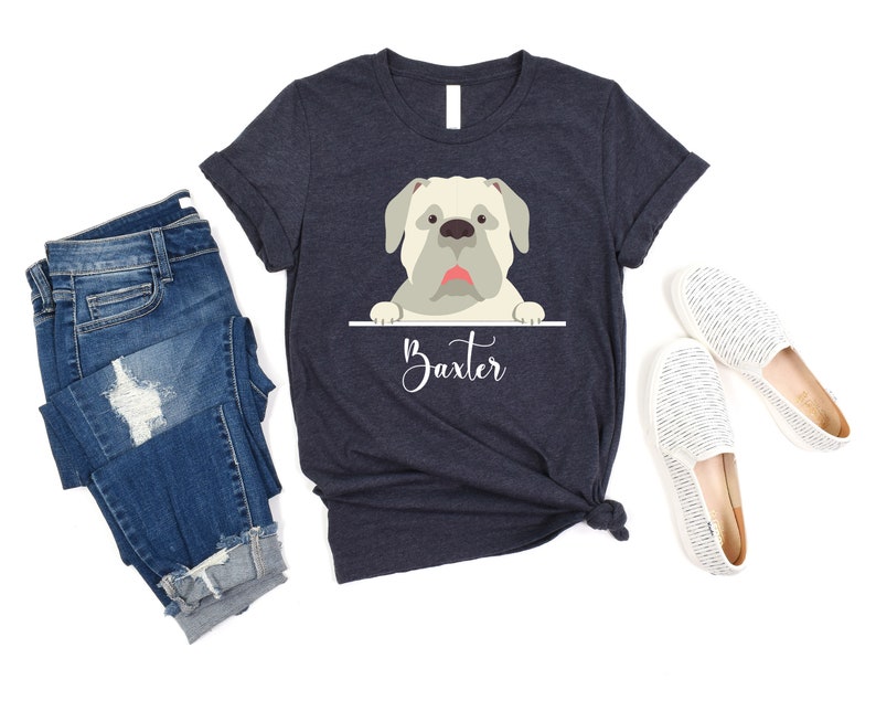 Peut inclure: Un t-shirt bleu marine avec une image de dessin anim&eacute; d'un chien blanc qui regarde par-dessus le haut d'une pancarte qui dit "Baxter".