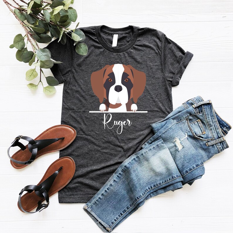 Peut inclure: Un t-shirt gris fonc&eacute; avec une illustration de dessin anim&eacute; d'un chien brun et blanc regardant par-dessus une barre avec le nom "Ruger" &eacute;crit en dessous. Le t-shirt est pos&eacute; sur une surface blanche avec une paire de sandales brunes et un jean bleu.