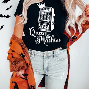 May include: A black t-shirt with a white graphic of a slot machine with the text "Jackpot Slots" and "Queen of the Machine".