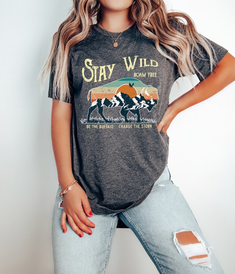 May include: A gray t-shirt with a vintage-style graphic of a buffalo walking in front of a mountain range with a sunset in the background. The text "Stay Wild" is above the buffalo and "Born Free" is below. The text "Be the Buffalo, Charge the Storm" is below the buffalo.