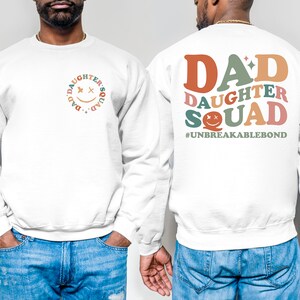 Puede incluir: Sudadera blanca con un gr&aacute;fico colorido en la espalda que dice "DAD DAUGHTER SQUAD #UNBREAKABLEBOND". La parte delantera de la sudadera tiene un gr&aacute;fico m&aacute;s peque&ntilde;o de una cara sonriente con las palabras "DAD'S DAUGHTER SQUAD" alrededor.
