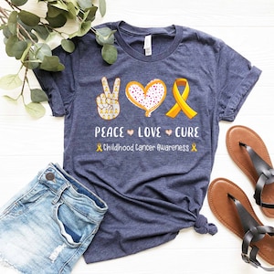May include: A navy blue t-shirt with a peace sign, heart, and yellow ribbon. The text "Peace Love Cure" and "Childhood Cancer Awareness" is printed on the shirt.