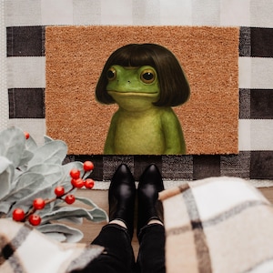 May include: A doormat featuring a green frog with a dark bob haircut. The frog is centered on a brown mat, set against a black and white checkered pattern. The image is taken from a first-person perspective, with black boots and a plaid blanket in view.