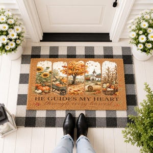 May include: A rectangular tan coir doormat featuring a multi-panel autumn scene with sunflowers, a wheelbarrow of pumpkins, a large maple tree, a church, and a basket of corn. The text reads HE GUIDES MY HEART through every harvest.