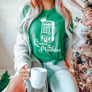 May include: A green t-shirt with a white graphic of a slot machine with the text "Jackpot Slots" and "Queen of the Machine".